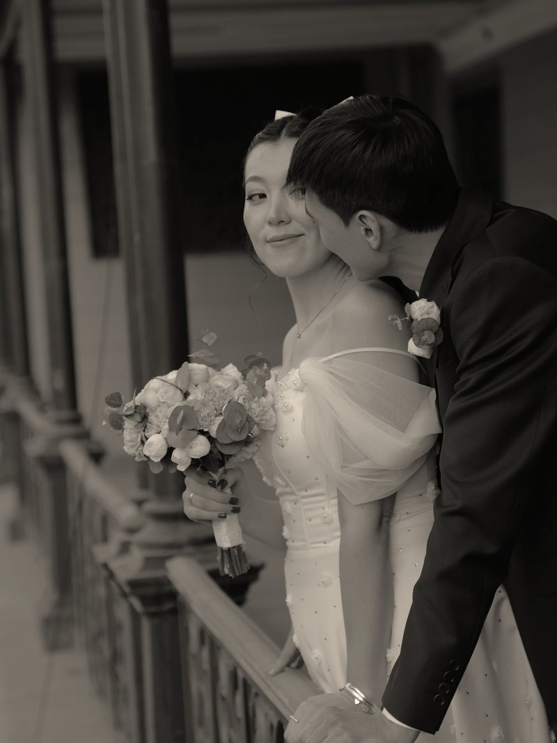 Groom kissing the bride's cheek as she leans on a balcony railing with her bouquet